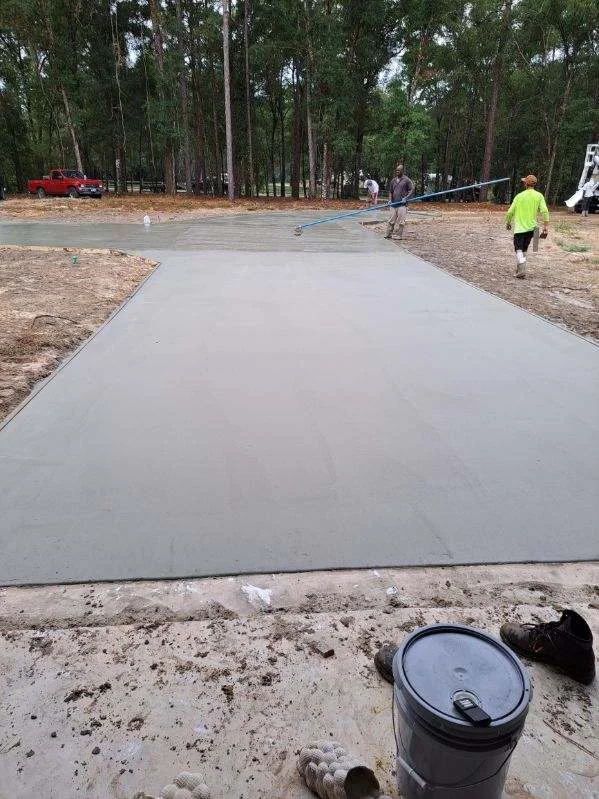 Newly poured concrete driveway with finishing work in progress, surrounded by pine trees