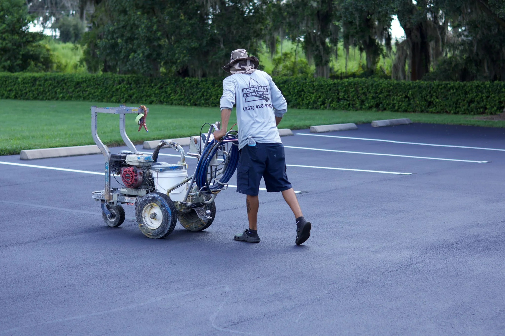 Worker operating sealcoating spray equipment on dark asphalt parking lot with white line markings