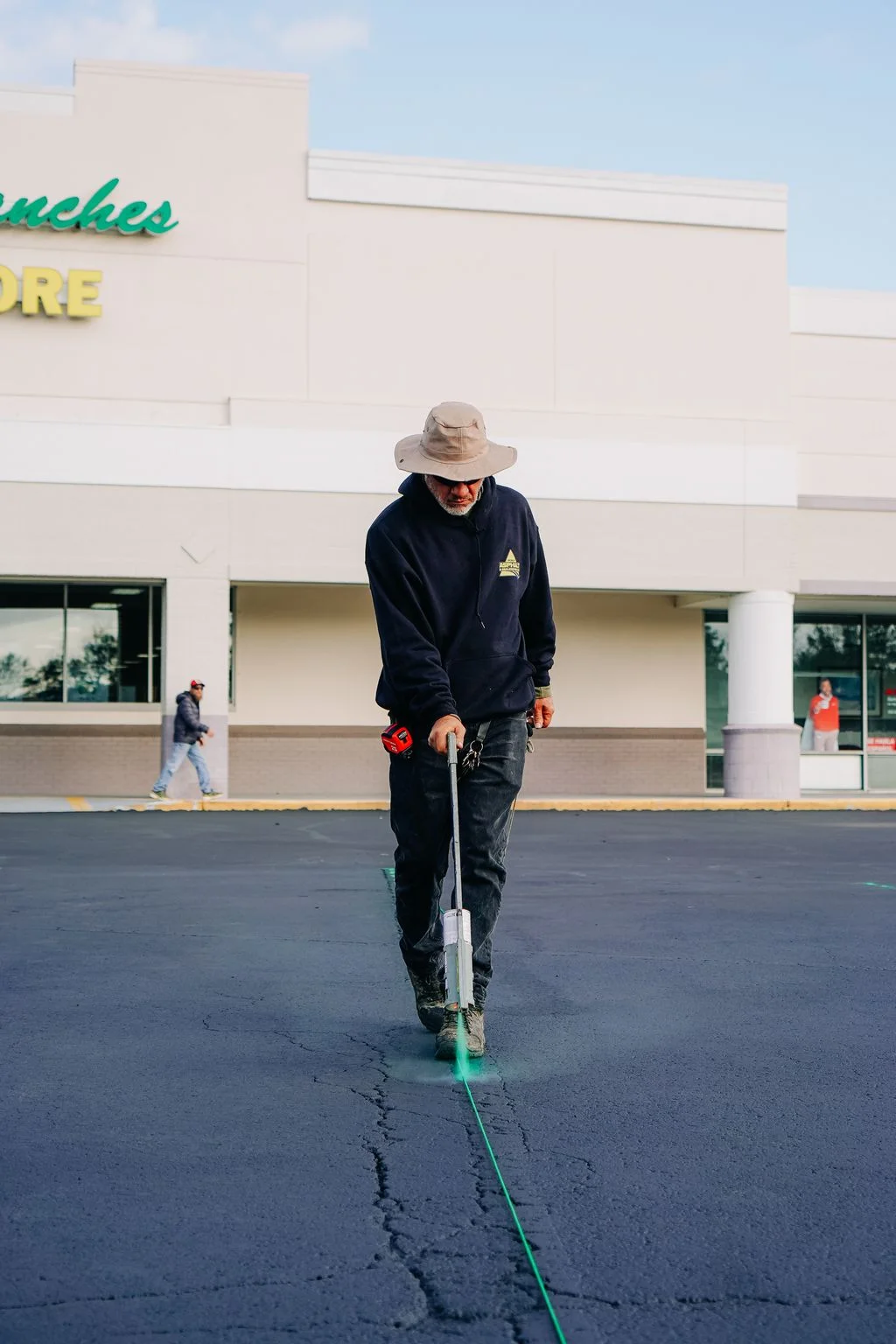 Technician applying green crack sealant to parking lot asphalt with commercial building in background